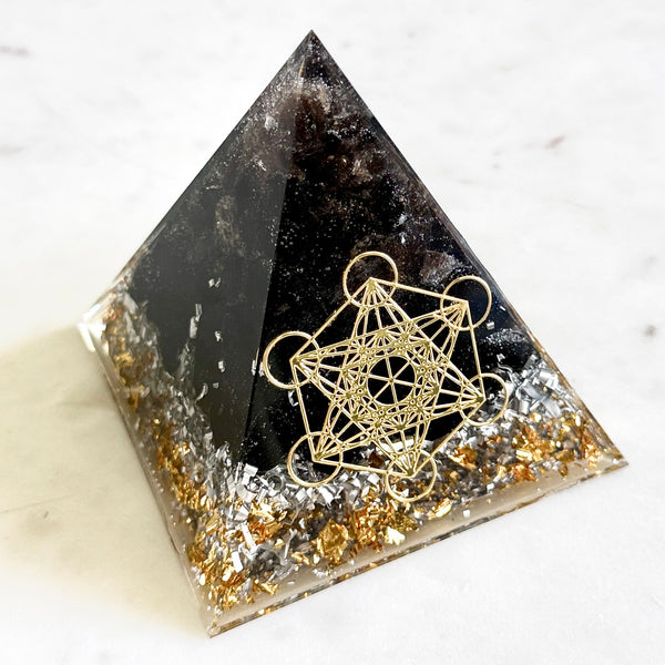 A clear resin pyramid with smoky quartz and black obsidian chips, embedded with metallic gold and silver flakes along the base, and featuring a gold geometric star and circle design on the front, set on a light marble surface.