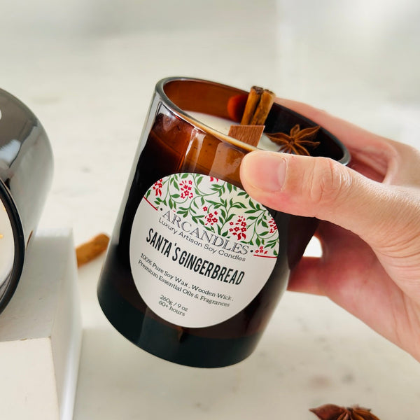 Hand holding a 'Santa's Gingerbread' candle with a decorative label on a light background