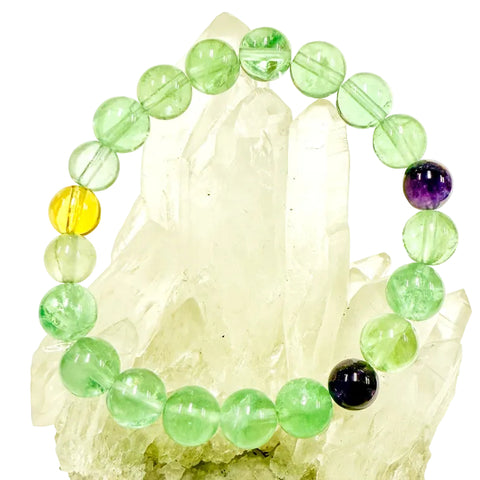 A bracelet made of round green and purple fluorite beads displayed on a crystal-like surface.